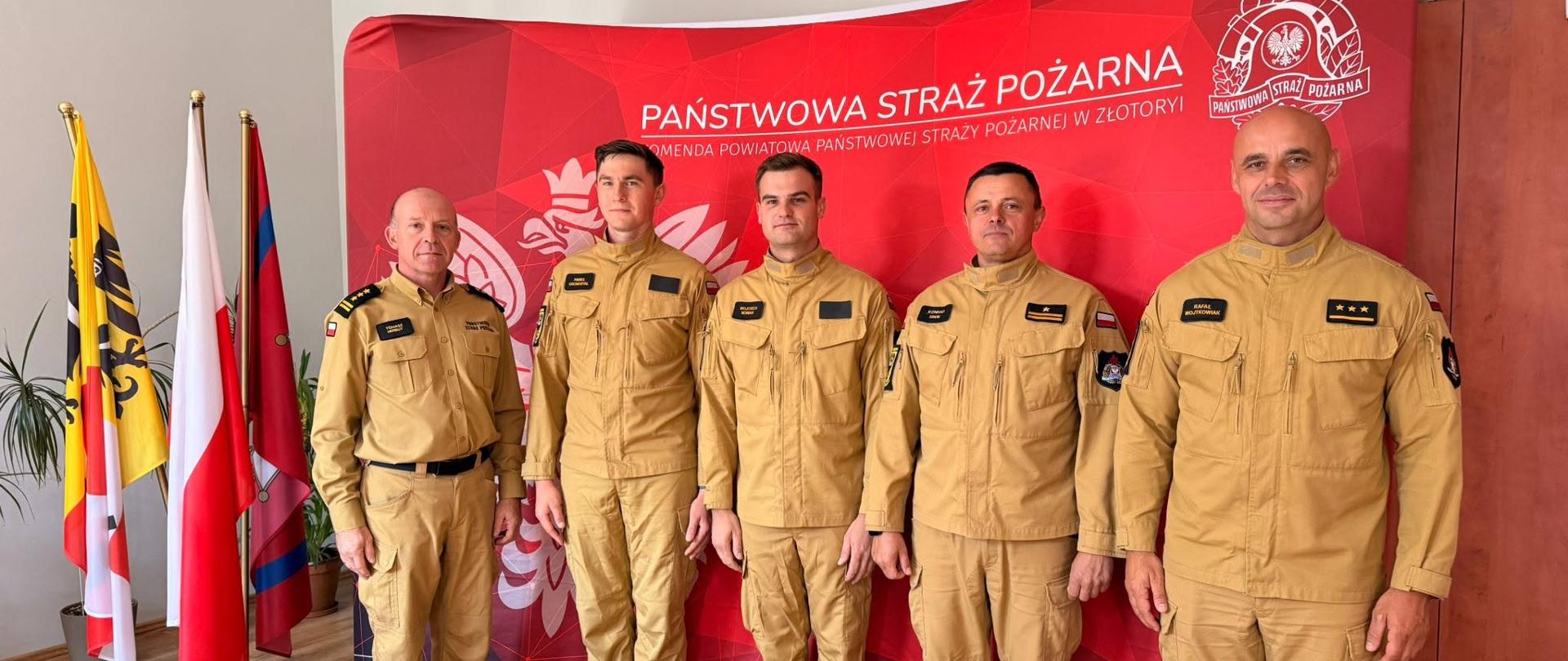 pięciu strażaków stoi w szeregu, umundurowanie służbowe. Biuro, w tle czerwony baner z logo PSP, obok stojak z trzema flagami