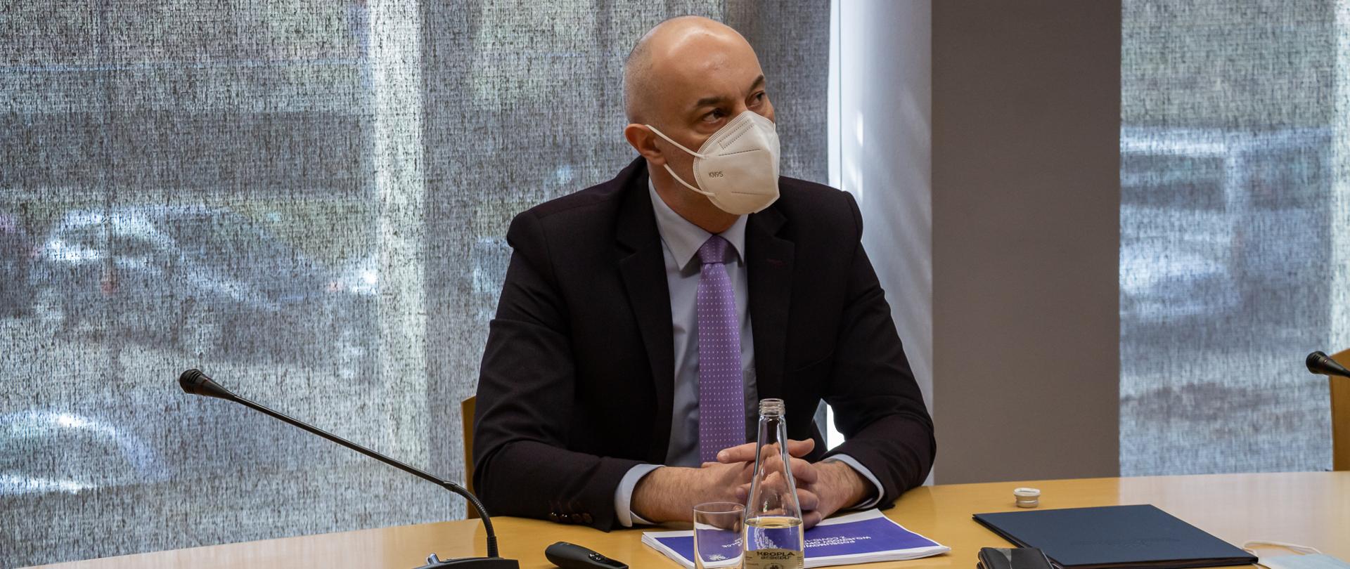 Na zdjęciu zasiadający za stołem wojewoda opolski Adrian Czubak, który referuje działania rządu na rzecz walki z epidemią koronawirusa, podczas posiedzenia WRDS w Opolu. Wojewoda patrzy ukosem w stronę monitora, ma spelcione słonie, przed nim na stole leżą dokumenty. Obok dokumentów szklana butelka z wodą oraz pusta szklanka. Na stole znajduje się również mikrofon bezprzewodowy, telefonu komórkowe oraz granatowa teczka. Z uwagi na stan epidemii, wojewoda na twarzy ma maseczkę ochronną. Wojewoda zasiada na tle zasłoniętego okna. 