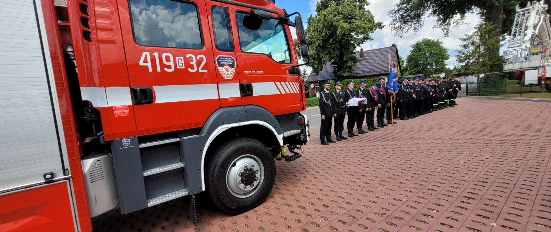 Wybrukowany plac na placu od lewej strony widoczny z boku przedni fragment pojazdu pożarniczego. W oddali strażacy w umundurowaniu galowym, zbiórka w dwuszeregu. 