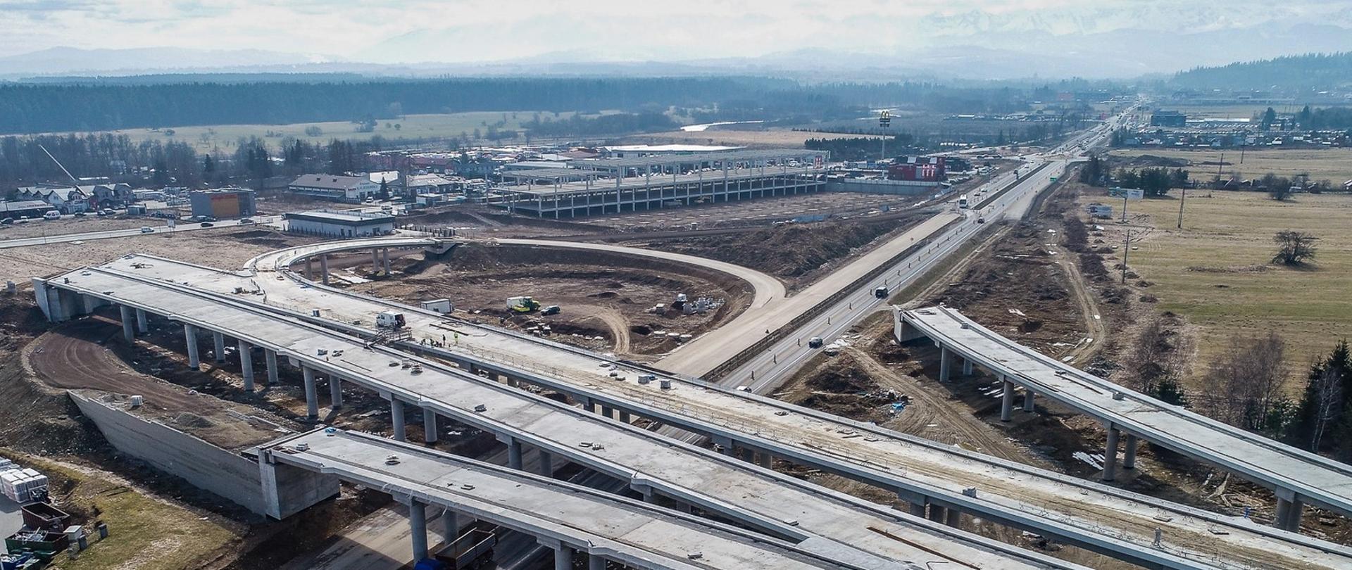 węzeł drogowy w trakcie budowy widziany z lotu ptaka, na drugim planie budowa hali, droga jednojezdniowa, w oddali ośnieżone szczyty gór