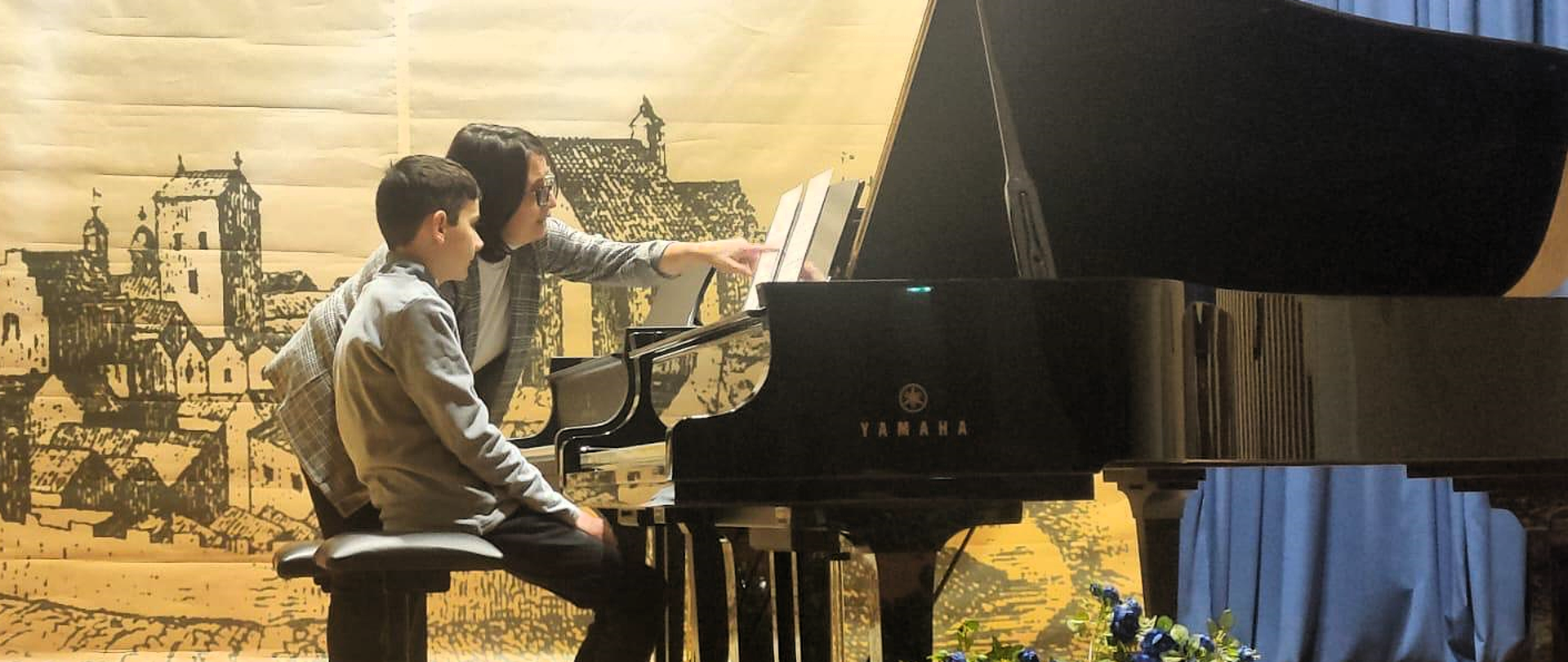 dziecko przy fortepianie, obok niego kobieta, za nimi beżowy baner