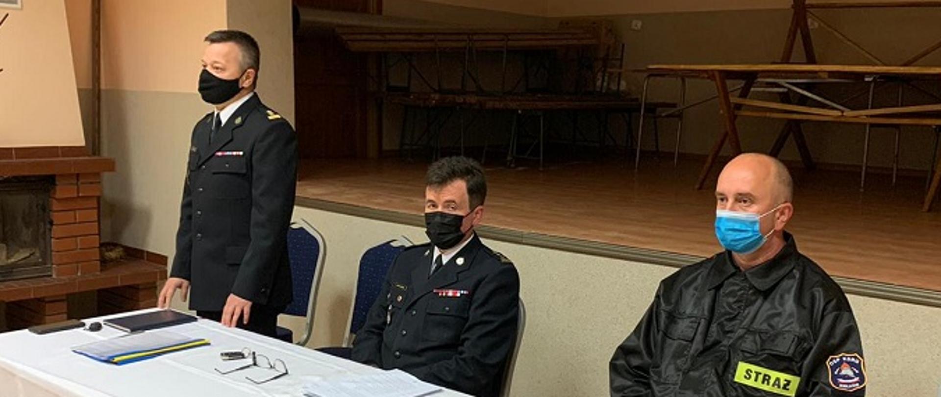 Zdjęcie zrobione w sali. Na fotografii przy stole prezydialny siedzą trzy osoby podczas spotkania. Wśród nich dwóch strażaków PSP ubranych w mundury wyjściowe oraz dh OSP ubrany w mundur koszarowy.