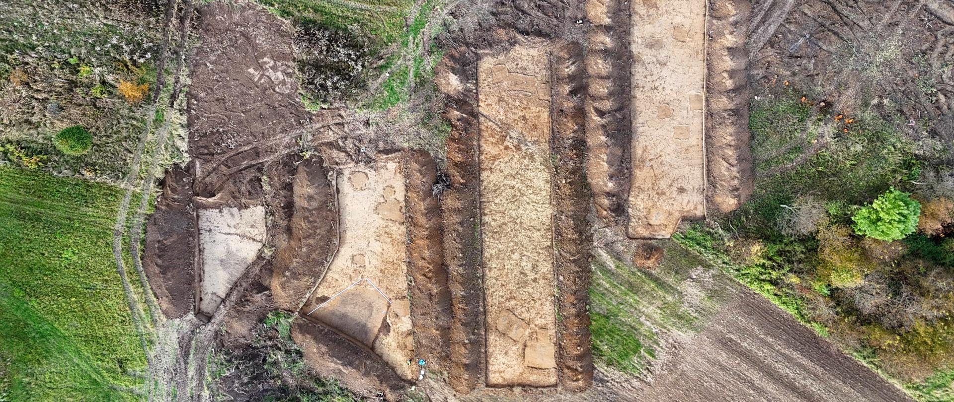 Na zdjęciu widać teren z lotu ptaka, prawdopodobnie pole lub łąkę, na którym znajdują się duże, prostokątne wykopy w ziemi. Jest ich kilka, ułożone są w rzędzie, a każdy ma wyraźnie odsłoniętą jasną warstwę gleby w środku, otoczoną ciemniejszą ziemią. Wokół wykopów widać ślady ciężkiego sprzętu – głębokie koleiny w błocie. Teren jest częściowo zielony, z fragmentami trawy i krzewów, a w prawym dolnym rogu widoczny jest kawałek zaoranego pola.