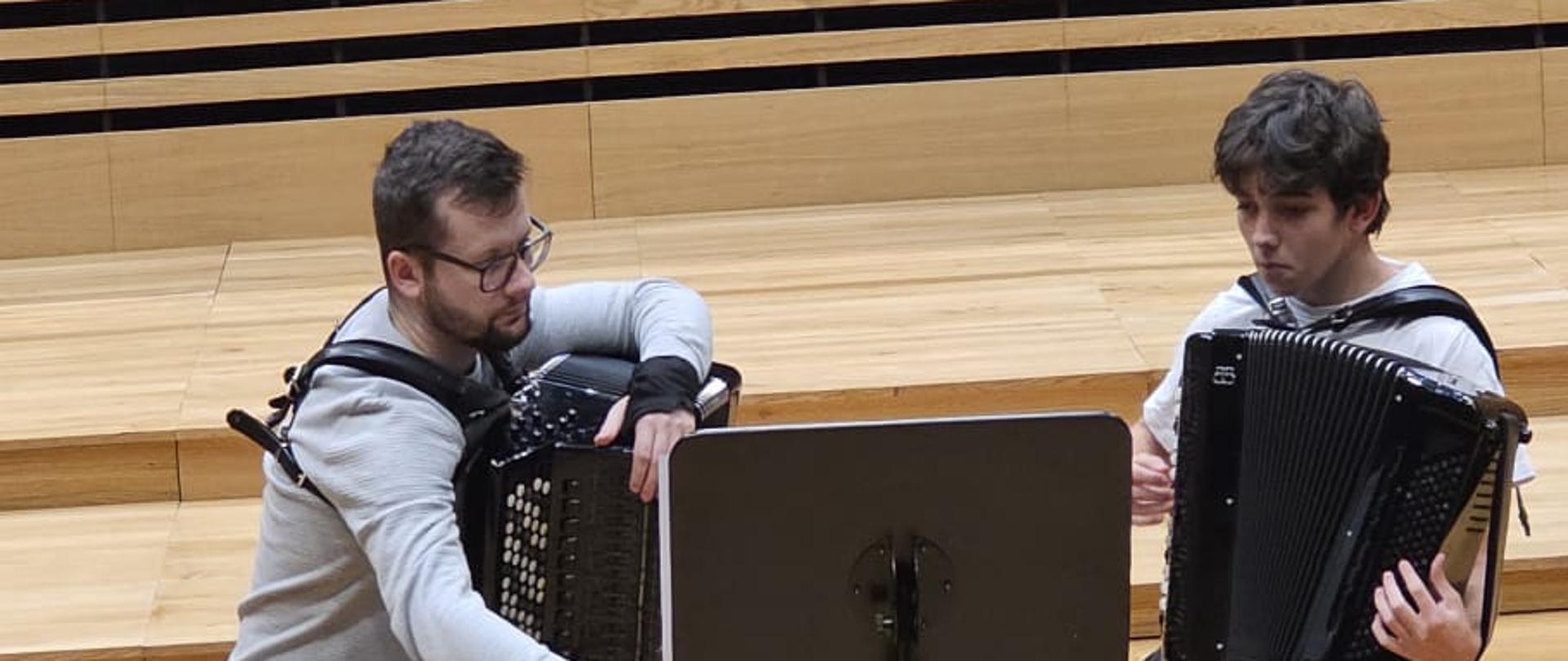 Na estradzie sali koncertowej PSM na krzesłach obok siebie siedzą mężczyzna i chłopak, przed nimi stoi pulpit. mężczyzna trzyma akordeon, chłopak gra na akordeonie.