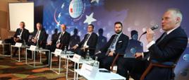 Widok na scenę, na której siedzą paneliści: od prawej: moderator dyskusji Jacek Żalek – sekretarz stanu w Ministerstwie Funduszy i Polityki Regionalnej, Grzegorz Rabsztyn z Europejskiego Banku Inwestycyjnego, Mariusz Strojny ze Szkoły Głównej Handlowej, Michał Góra – Prezes Alfavox, Wojciech Kamieniecki – dyrektor NCBR, Michał Janasik – Wiceprezes ds. Finansów i Komercjalizacji Centrum Łukasiewicz. Za panelistami plansza z logo i napisem: Economic Forum