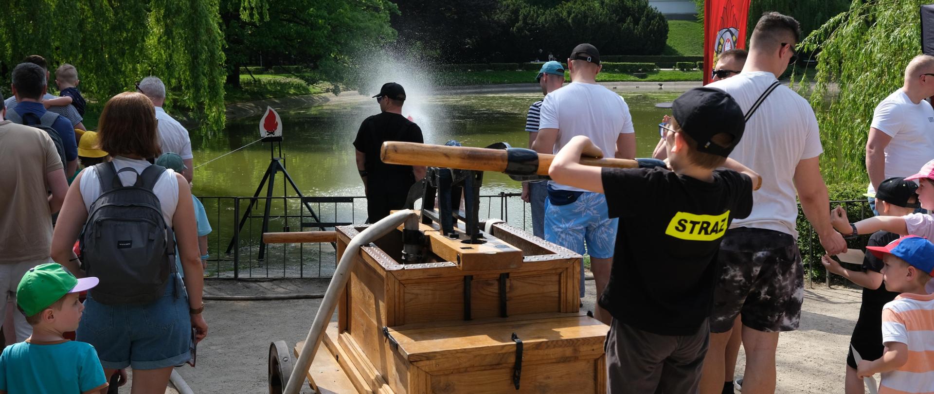 Dzieci pompują sikawką ręczną wodę podczas konkurencji celności trafienia prądem wody w tarczę.
