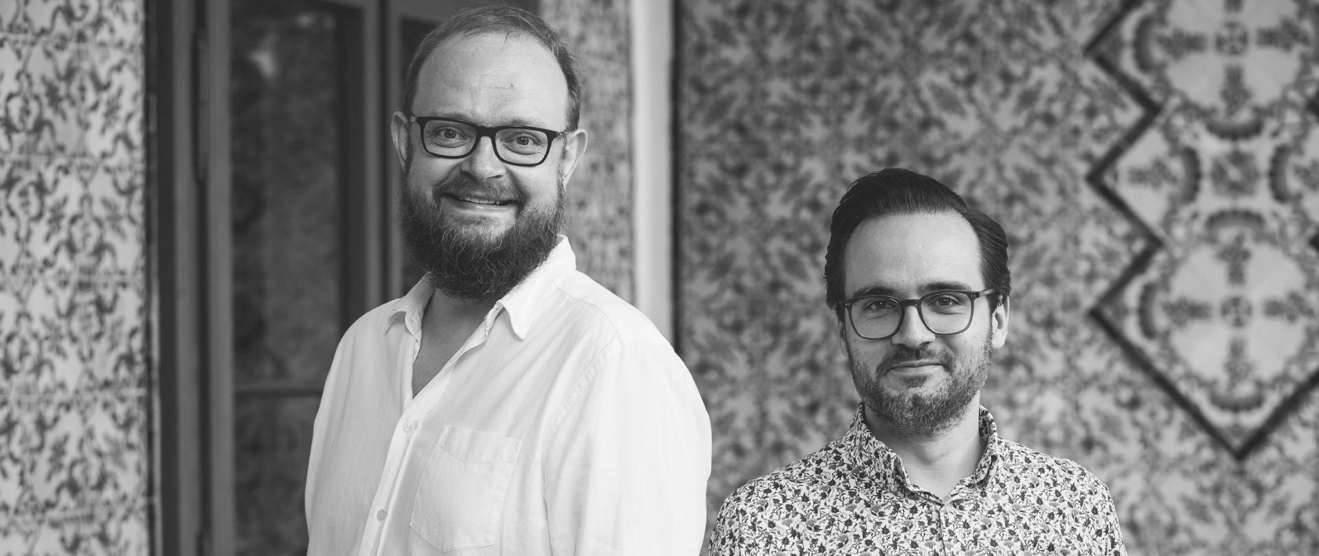 Dwóch mężczyzn – artyści fortepianowego duetu – stoi obok siebie na tle dekoracyjnej ściany. Oboje uśmiechają się do aparatu; pierwszy z lewej ma jasną koszulę i okulary, drugi ma ciemniejsze włosy, krótszą brodę oraz wzorzystą koszulę. To portret współczesnych pianistów z zespołu Zarębski Piano Duo, specjalizującego się w utworach na cztery ręce.