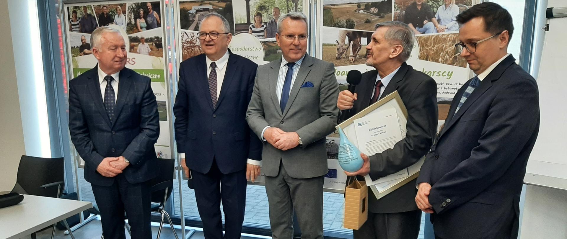 Grzegorz Smytry, dyrektor RZGW w Bydgoszczy dziękuje za współpracę. Stoi z mikrofonem w ręku. Trzyma również nagrody. Wypowiada się. Obok niego przedstawiciele innych instytucji. 