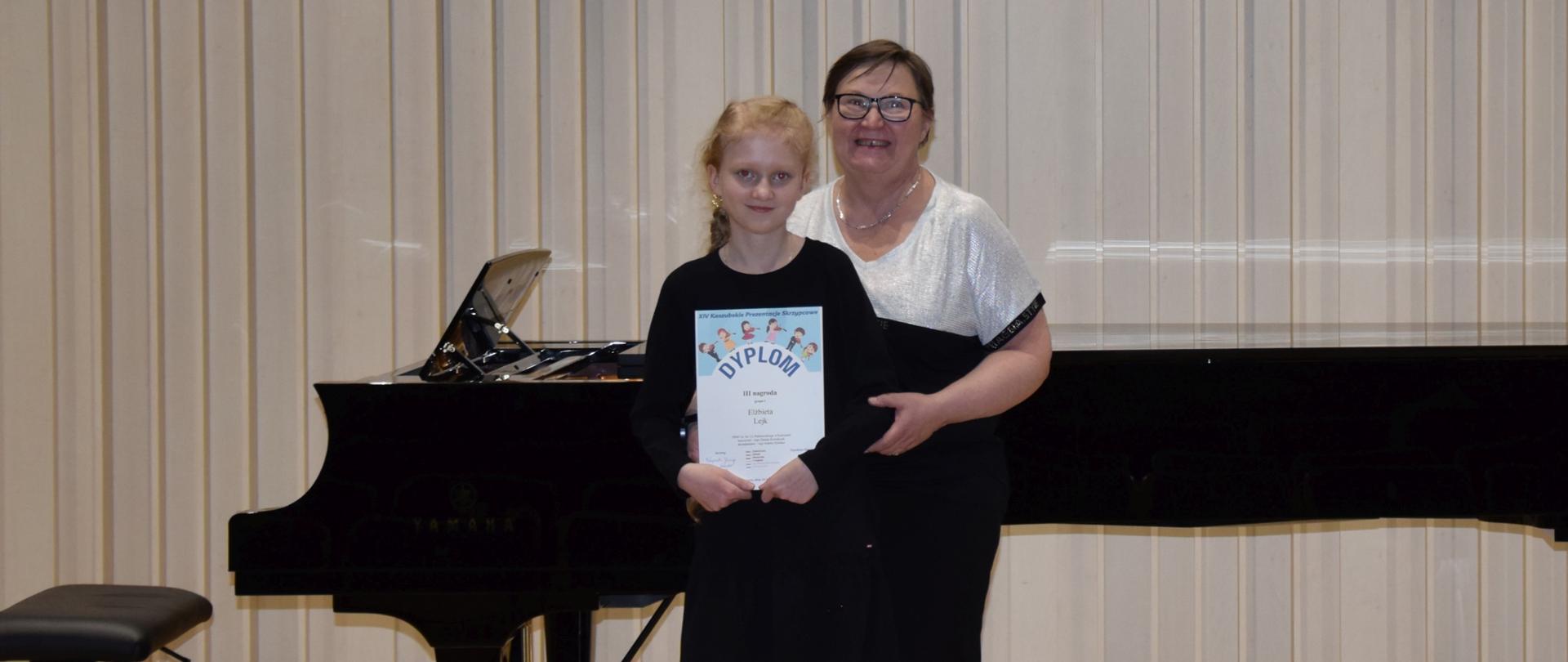 Na scenie znajduje się uczennica trzymająca w rękach dyplom wraz z nauczycielką