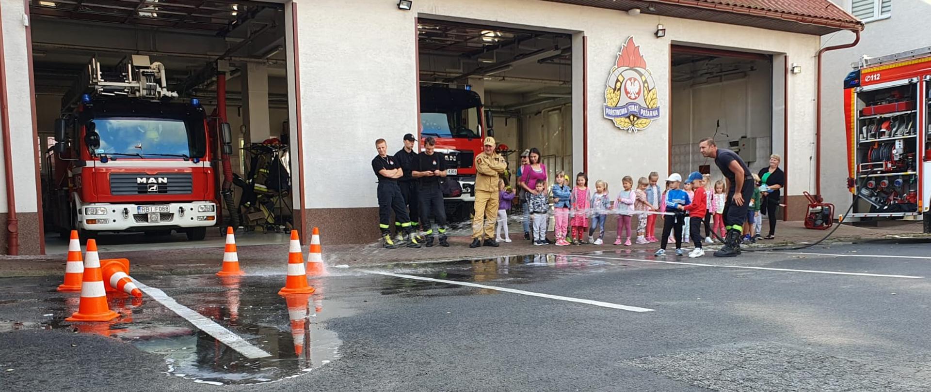 Zdjęcie przedstawia dzieci które próbują strącić słupki przy użyciu szybkiego natarcia oraz strażaków którzy stoją przy nich. Widać również samochody pożarnicze które stoją w garażach Komendy Powiatowej PSP w Ustrzykach Dolnych. 