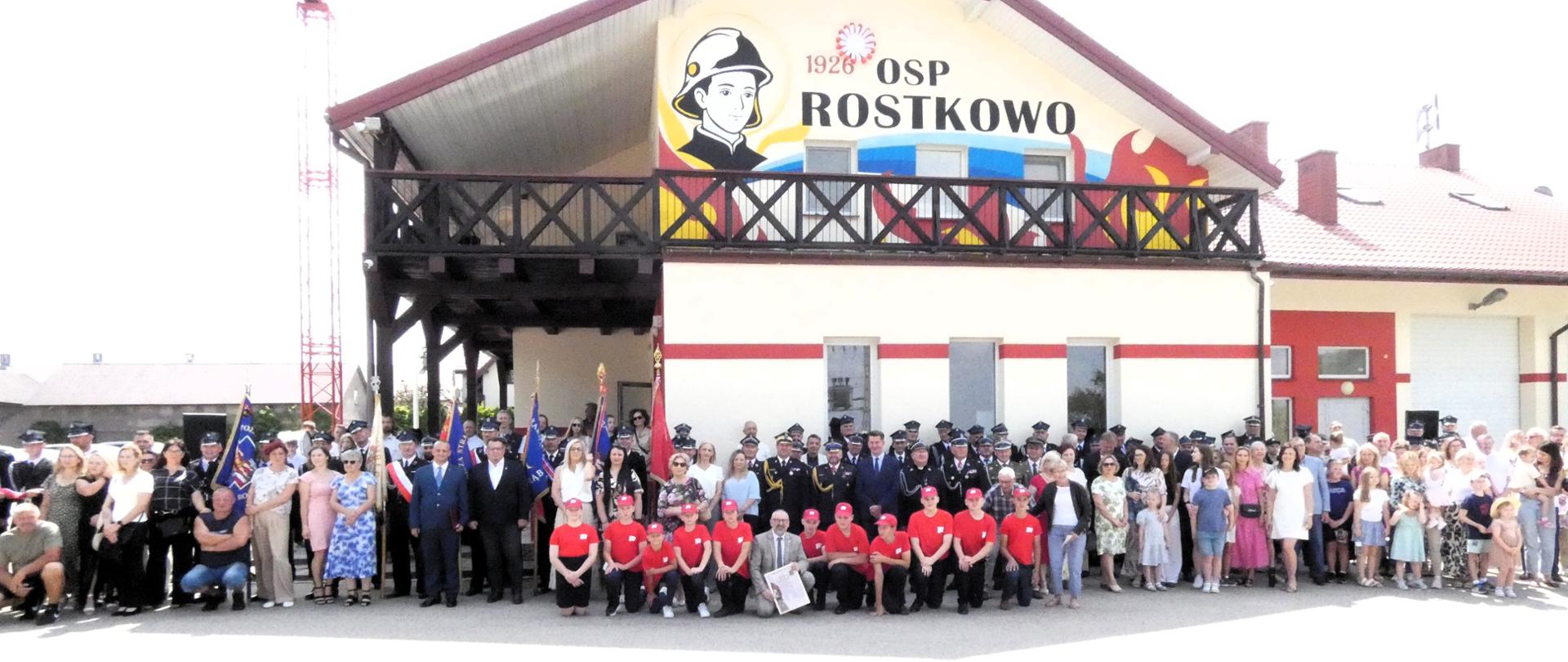 Zdjęcie grupowe uczestników wydarzenia przed budynkiem, którego szczyt zdobi mural "1926 OSP ROSTKOWO". 