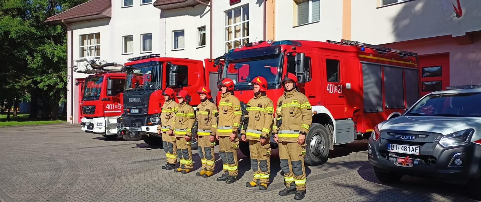 na zdjęciu widać stojących strażaków w tle samochody strażackie i budynek KP PSP w A-stowie