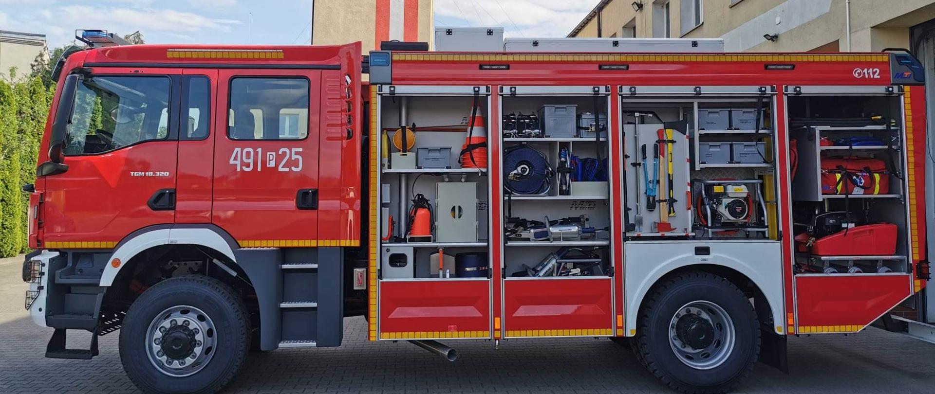 Zdjęcie przedstawia lewy bok nowego ciężkiego pojazdu ratowniczo - gaśniczego marki Man. W samochodzie otwarto skrytki sprzętowe uwidaczniając sprzęt znajdujący się na wyposażeniu auta. 