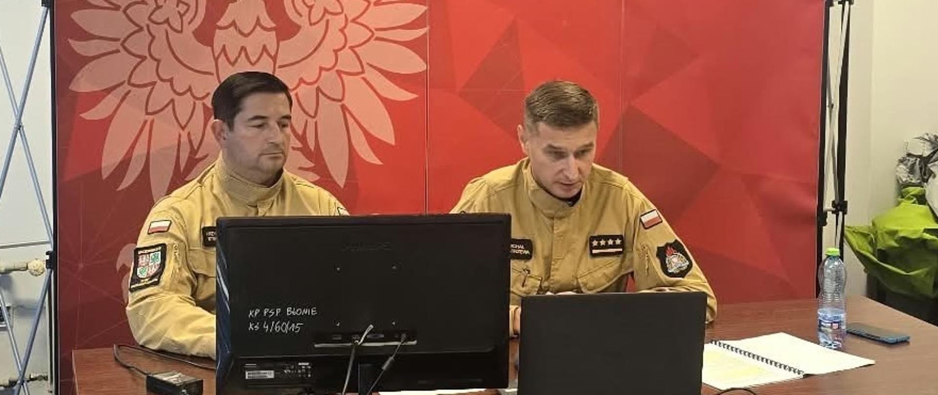 Na zdjęciu przedstawionych jest dwóch strażaków Państwowej Straży Pożarnej (PSP) siedzących przy długim, drewnianym stole w pomieszczeniu biurowym. Oto szczegółowy opis tego, co znajduje się na fotografii:Osoby na zdjęciu:W pierwszym planie siedzi strażak w piaskowym mundurze służbowym PSP z polską flagą oraz naszywką ze stopniem (cztery gwiazdki – kapitan). Posiada również imiennik (z nazwiskiem Kostrzewa). Operuje myszką podłączoną do laptopa. Za nim siedzi drugi strażak, również w piaskowym mundurze służbowym PSP, który pisze coś na kartce papieru. Wyposażenie stołu:Otwarty laptop, monitor oraz myszka. Dokumenty, notatnik w oprawie spiralnej oraz długopis. Butelka wody mineralnej, smartfon oraz drobne akcesoria. Tło i otoczenie:Po prawej stronie stoi duży, czerwony ścianka/baner z godłem Polski (orłem w koronie) oraz napisem „PAŃSTWOWA STRAŻ POŻARNA”. Na dole baneru widnieje napis dotyczący Komendy Powiatowej Państwowej Straży Pożarnej w Powiecie Warszawskim Zachodnim z siedzibą w Ożarowie Mazowieckim (ul. Lesznowska 20 A) oraz dane kontaktowe. Po lewej stronie widać okna przesłonięte niebieskimi pionowymi żaluzjami (verticalami). Podłoga wyłożona jest ciemną wykładziną dywanową. Sytuacja wygląda na prowadzenie szkolenia online, odprawy, konferencji prasowej lub spotkania w formie wideokonferencji. 