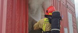 strażak gaszący pożar metalowego kontenera z którego wydobywa się gęsty szary dym