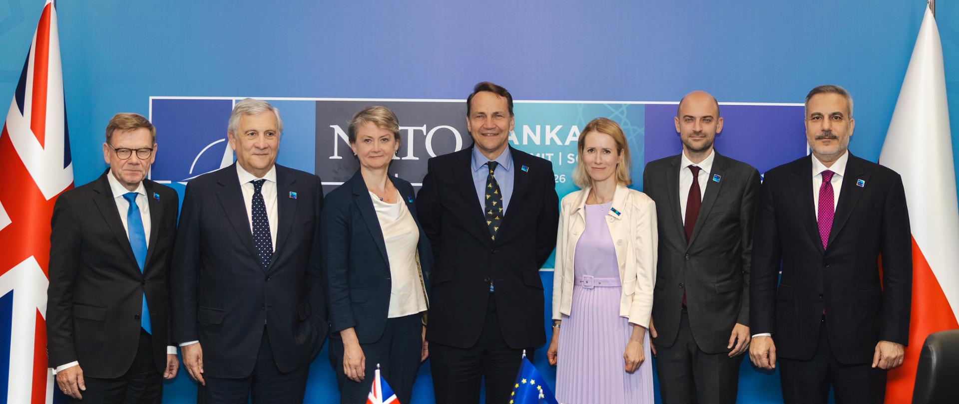 Siedem osób pozuje do wspólnego zdjęcia podczas spotkania międzynarodowego. Po środku stoi Wicepremier Sikorski. Przed grupą znajduje się stół konferencyjny z małymi flagami Wielkiej Brytanii, Francji i Unii Europejskiej, butelkami wody, szklankami i dokumentami. W tle widoczne są flagi Wielkiej Brytanii i Polski oraz niebieska ścianka z logo szczytu NATO.