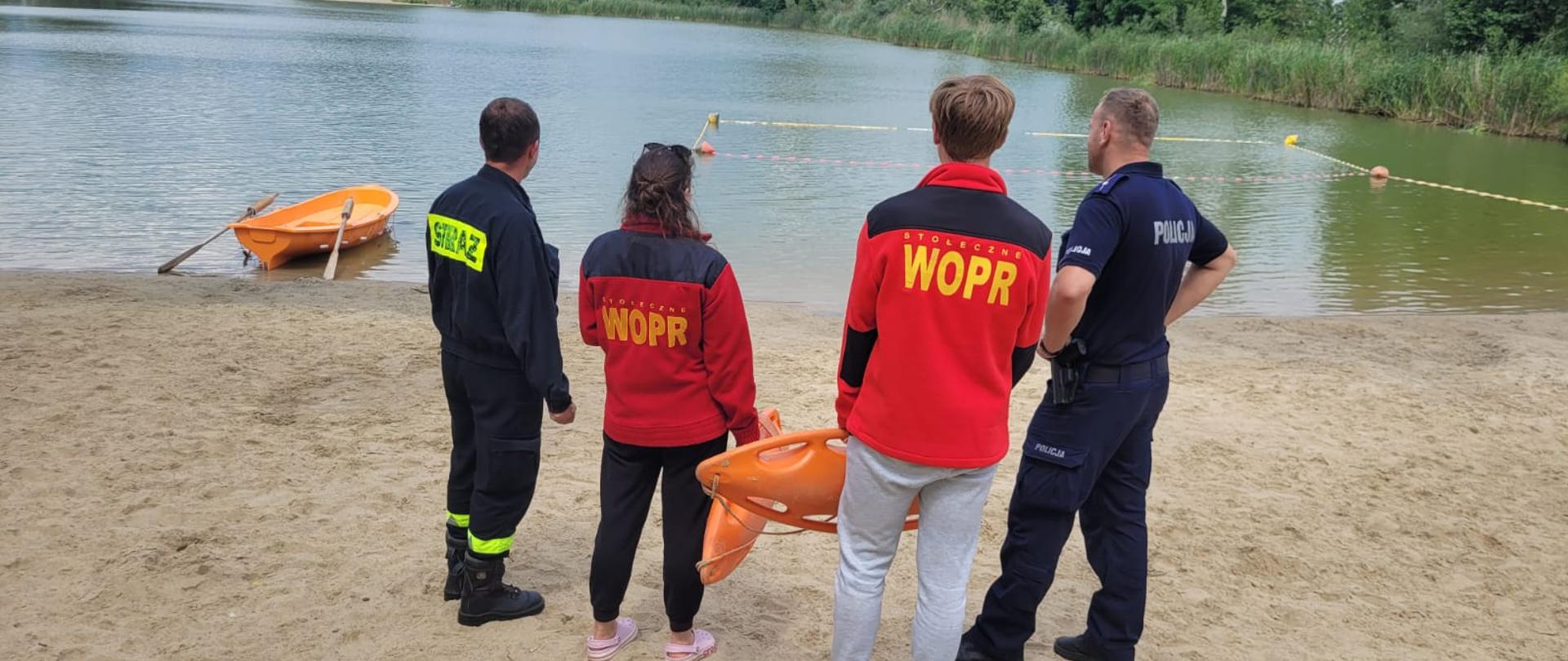 Zdjęcie przedstawia patrol składający się ze strażaka oraz policjanta. Funkcjonariusze stoją na piaszczystej plaży, rozmawiają z ratownikami wopr. W tle widać akwen zielonkowskich glinianek oraz wyznaczone bojkami kąpielisko.
