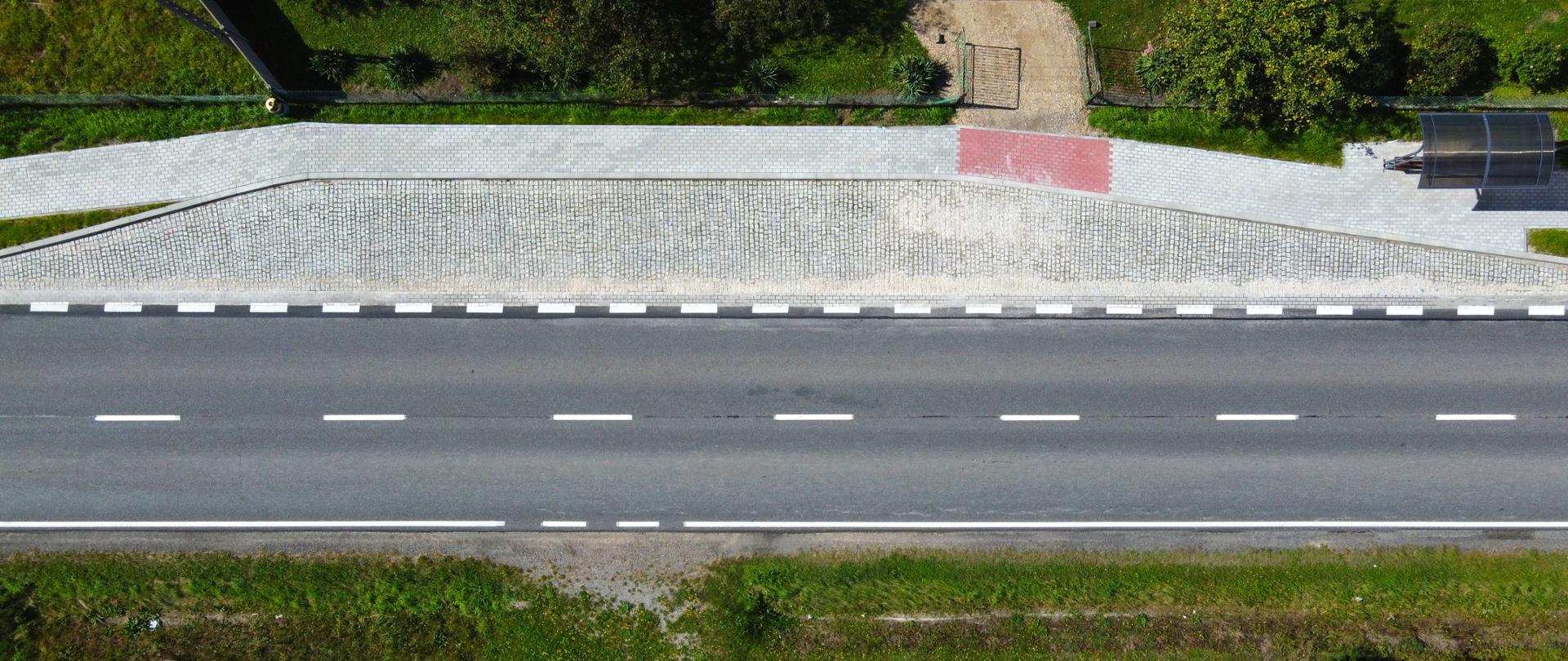 Widok z lotu ptaka na prosty odcinek asfaltowej drogi. Po jednej stronie jezdni znajduje się szeroki chodnik z kostki brukowej, zatoka autobusowa z wiatą oraz zjazd na posesję. Po obu stronach drogi widoczna jest zieleń: trawniki, krzewy i drzewa. Zdjęcie wykonano w słoneczny dzień, bez widocznych pojazdów i pieszych.