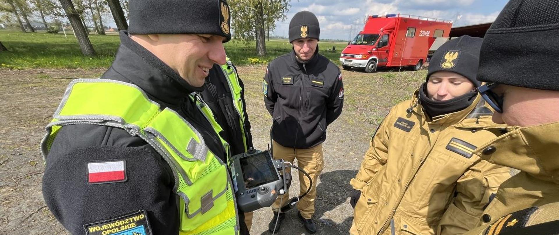 Ćwiczenia doskonalące dla pilotów bezzałogowych statków powietrznych Państwowej Straży Pożarnej województwa opolskiego. Piloci w umundurowaniu służbowym oraz w kamizelkach odblaskowych obsługują kontrolery bezzałogowych statków powietrznych podczas ćwiczeń. W tle widoczne drzewa oraz samochód dowodzenia i łączności Komendy Wojewódzkiego PSP w Opolu.