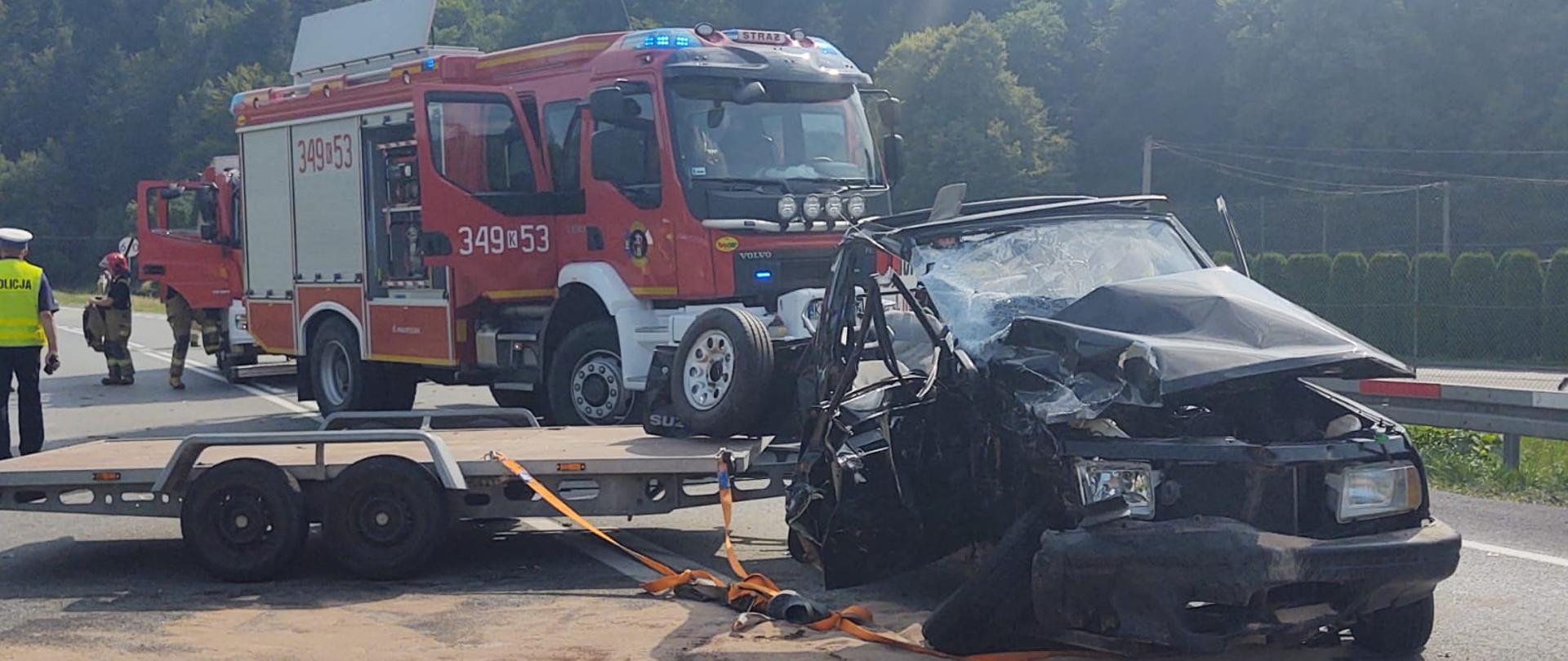 Samochód osobowy z przyczepą na drodze. Na drodze sorbent. W tle samochód pożarniczy.