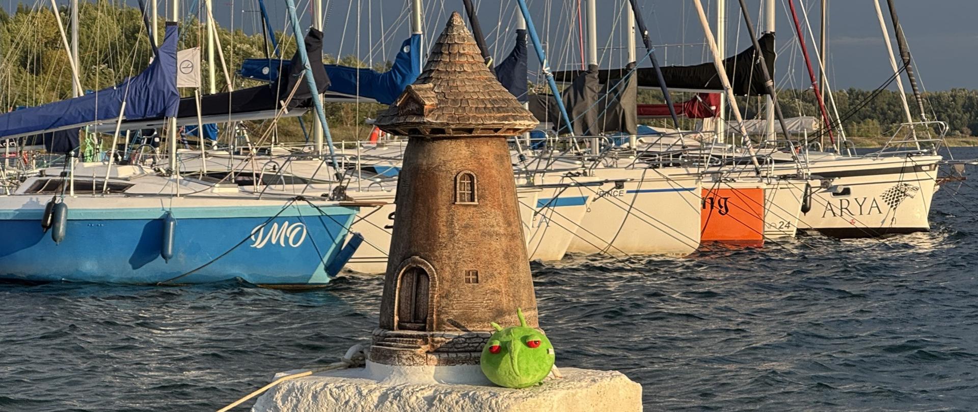 Przystań z zacumowanymi jachtami i żaglówkami przy drewnianym pomoście. Na pierwszym planie piaszczysty brzeg z kamieniami, a na końcu falochronu stoi mała dekoracyjna latarnia przy której siedzi zielona maskotka. Woda jeziora jest przejrzysta, a niebo częściowo zachmurzone, oświetlone promieniami popołudniowego słońca.