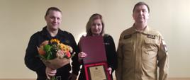 Komendant Powiatowy w Grodzisku Mazowieckim st. bryg. Krzysztof Tryniszewski wraz ze swoim zastępcą st. bryg. Piotrem Księżakiem wręczają kwiaty i pamiątkowy dyplom mł. kpt. Marioli Piątkowskiej odchodzącej na emeryturę. 