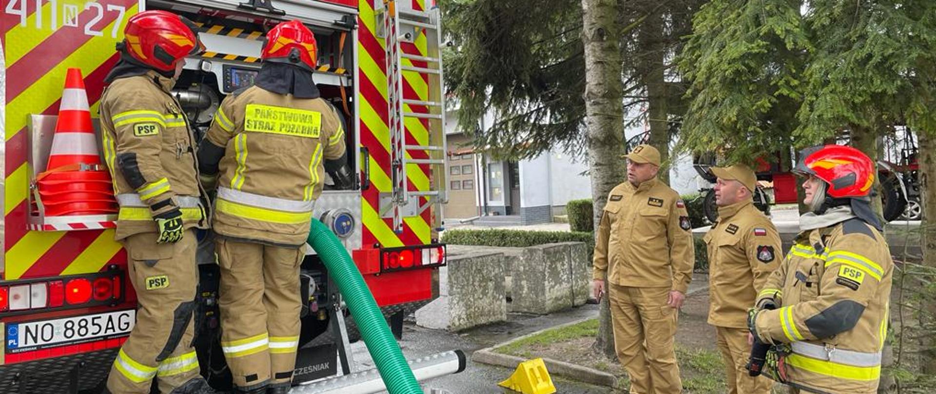 z lewej tył samochodu strażackiego, strażacy obsługują pompę pożarniczą