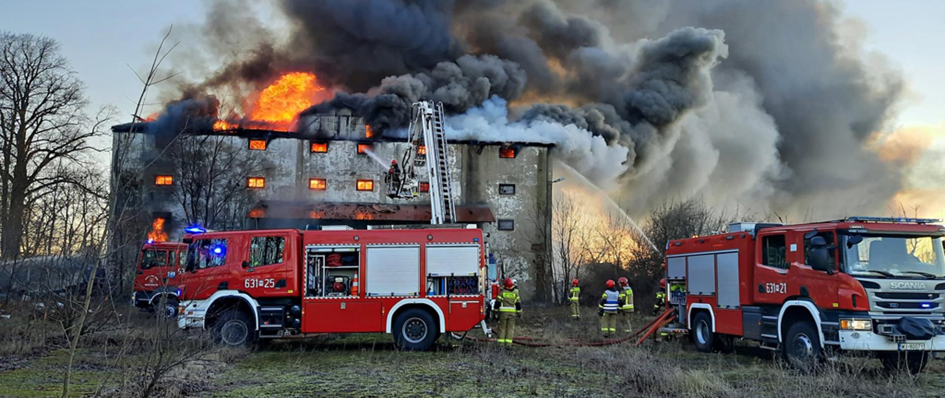Na zdjęciu widoczny budynek po dawnym spichlerzu w całości objęty pożarem. Nad dachem widoczny ogień i chmura czarnego dymu. Przed budynkiem swoją dwa samochody strażackie i kilku strażaków. Widoczne ramie podnośnika hydraulicznego, z kosza którego strażacy również gaszą pożar.