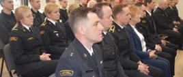 Grupa strażaków ubranych w mundury wyjściowe siedzi podczas narady, na pierwszym planie strażak w stopniu młodszego kapitana z naszywką Państwowa Straż Pożarna na prawym ramieniu