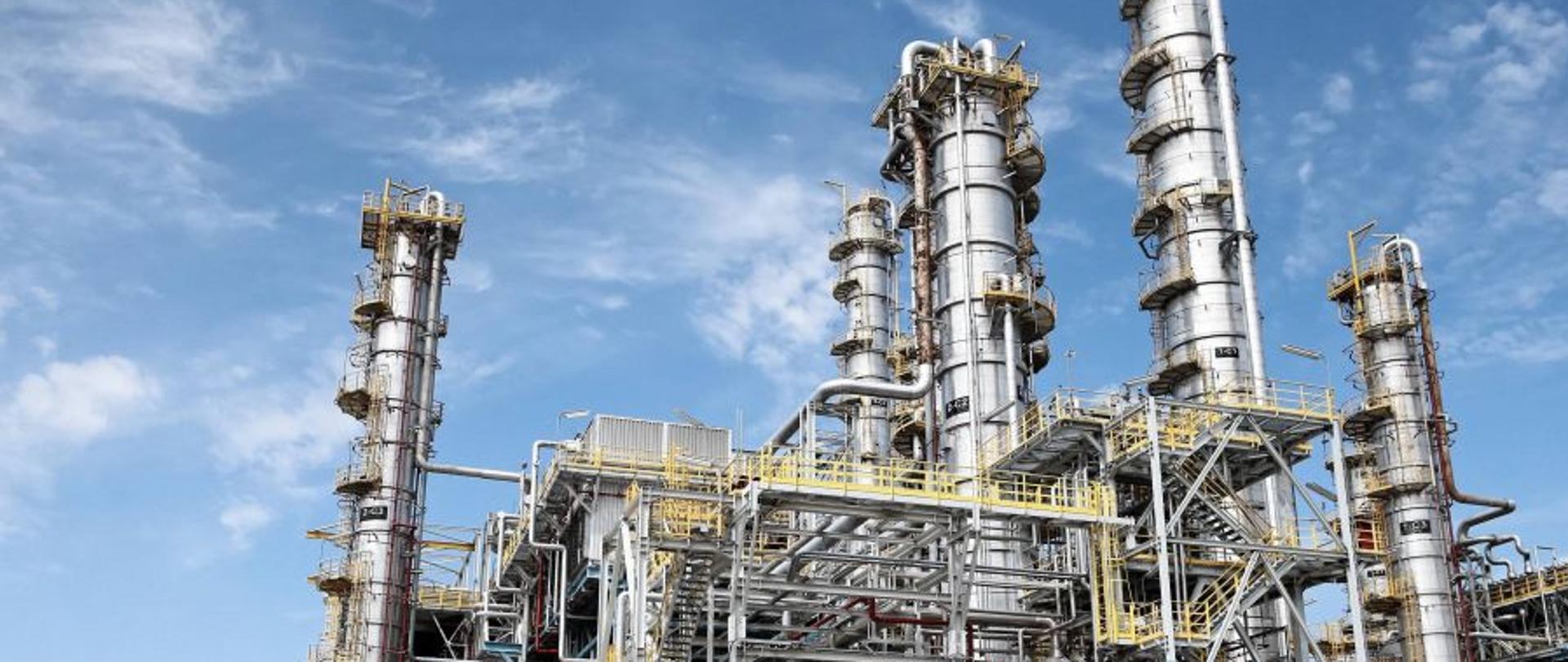 Duża instalacja przemysłowa: pięć okrągłych, wysokich na kilkanaście metrów szarych stalowych wież stojących na tle niebieskiego nieba pokrytego niewielkimi chmurami. Pomiędzy nimi widać gęstą sieć rurociągów i stalowe konstrukcje techniczne. 