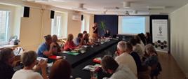 Uczestnicy narady siedzą przy stołach ustawionych w podkowę i słuchają prelegentki, która stoi przed ekranem z wyświetloną prezentacją. Obok niej przy stole siedzi druga osoba obsługująca prezentację. Na stołach znajdują się materiały szkoleniowe, notatniki oraz woda w dzbankach i szklanki. Po prawej stronie widoczny jest roll-up Państwowej Inspekcji Ochrony Roślin i Nasiennictwa. Wnętrze jest jasne, z dużymi oknami zapewniającymi naturalne oświetlenie.
