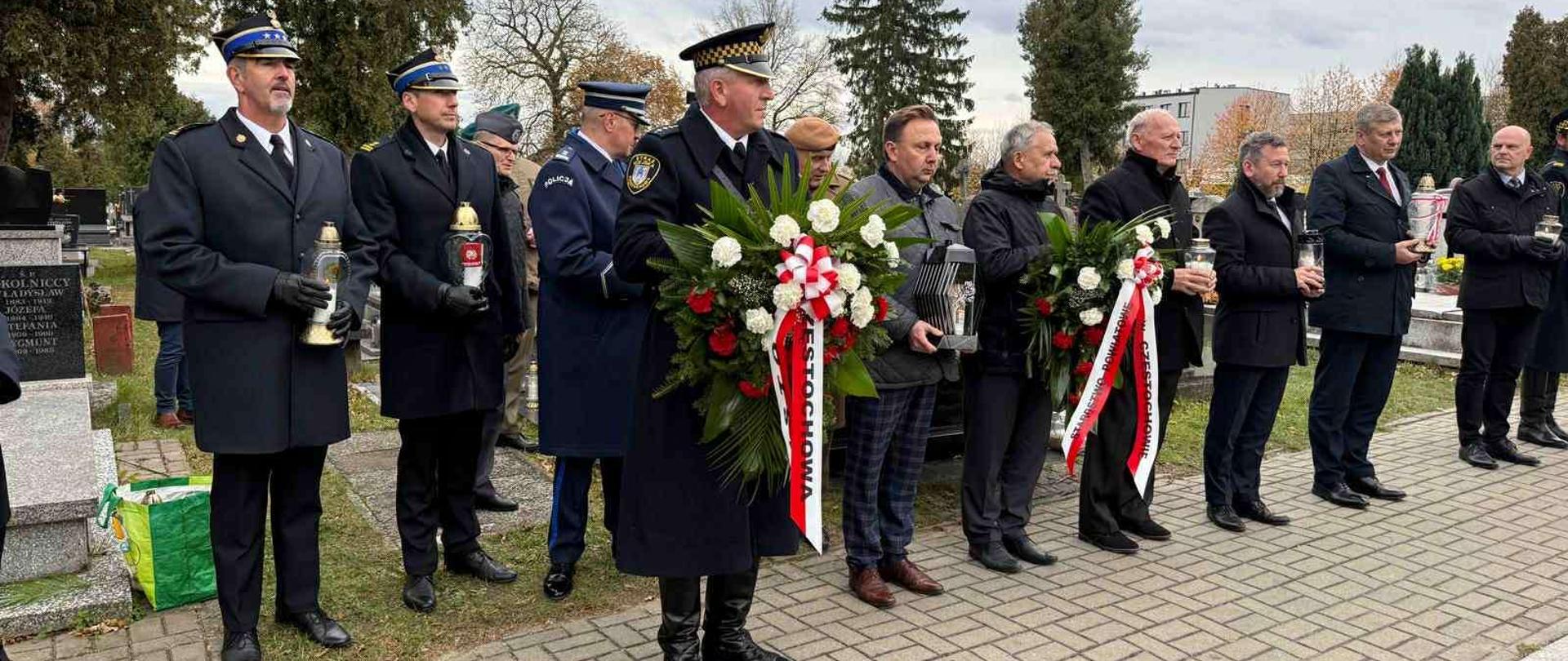 Przedstawiciele władz samorządowych oraz służb mundurowych, trzymający w dłoniach znicze bądź wieńce, stojący w alei cmentarnej przed grobami