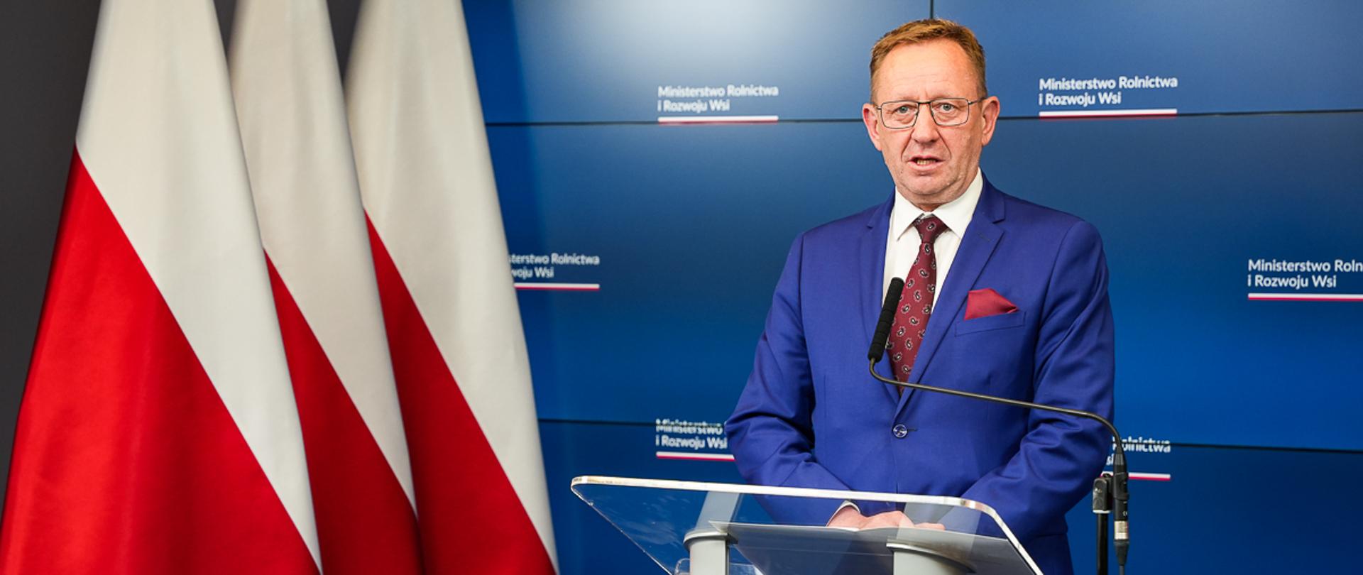 Minister Robert Telus stoi przy mównicy, w tle granatowy ekran z logo ministerstwa rolnictwa.