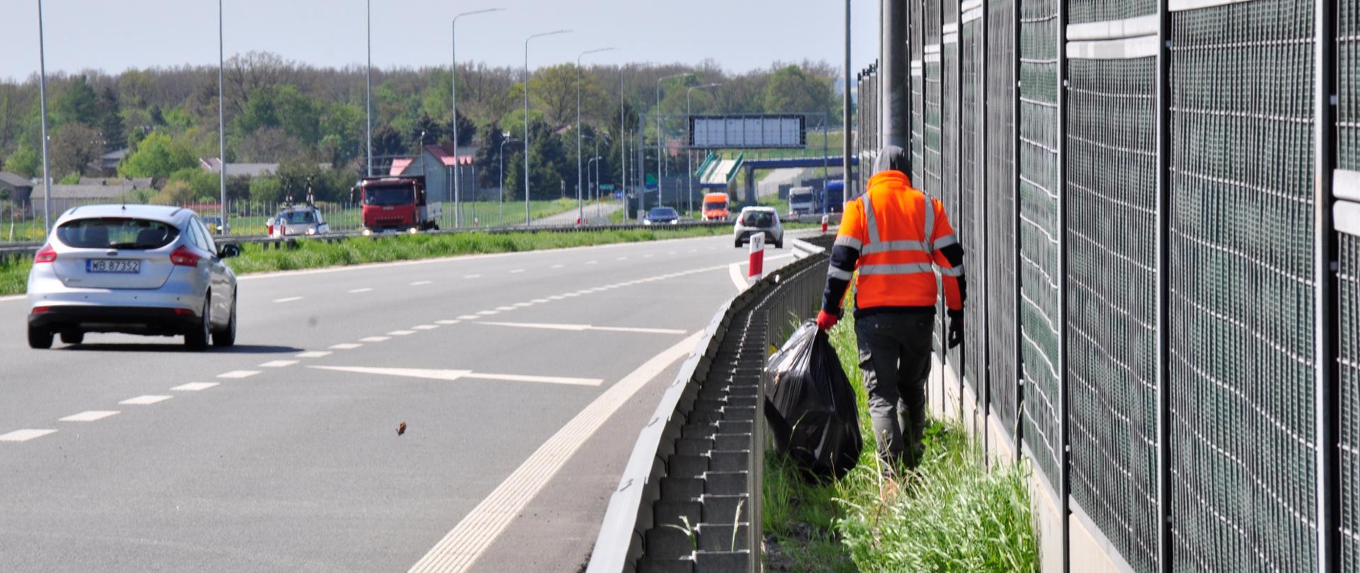 Zdjęcie przedstawia osobę ubraną w pomarańczową kamizelkę odblaskową, która porusza się wzdłuż pobocza autostrady, zbierając śmieci. Osoba ta trzyma duży czarny worek na śmieci i idzie obok metalowego ogrodzenia dźwiękochłonnego. Na drodze w tle widoczne są pojazdy, w tym srebrny samochód osobowy oraz czerwona ciężarówka. Po lewej stronie zdjęcia znajduje się pas zieleni oddzielający jezdnię od pobocza. W tle widoczne są drzewa oraz budynki, a dzień jest pogodny, z lekkim zachmurzeniem.