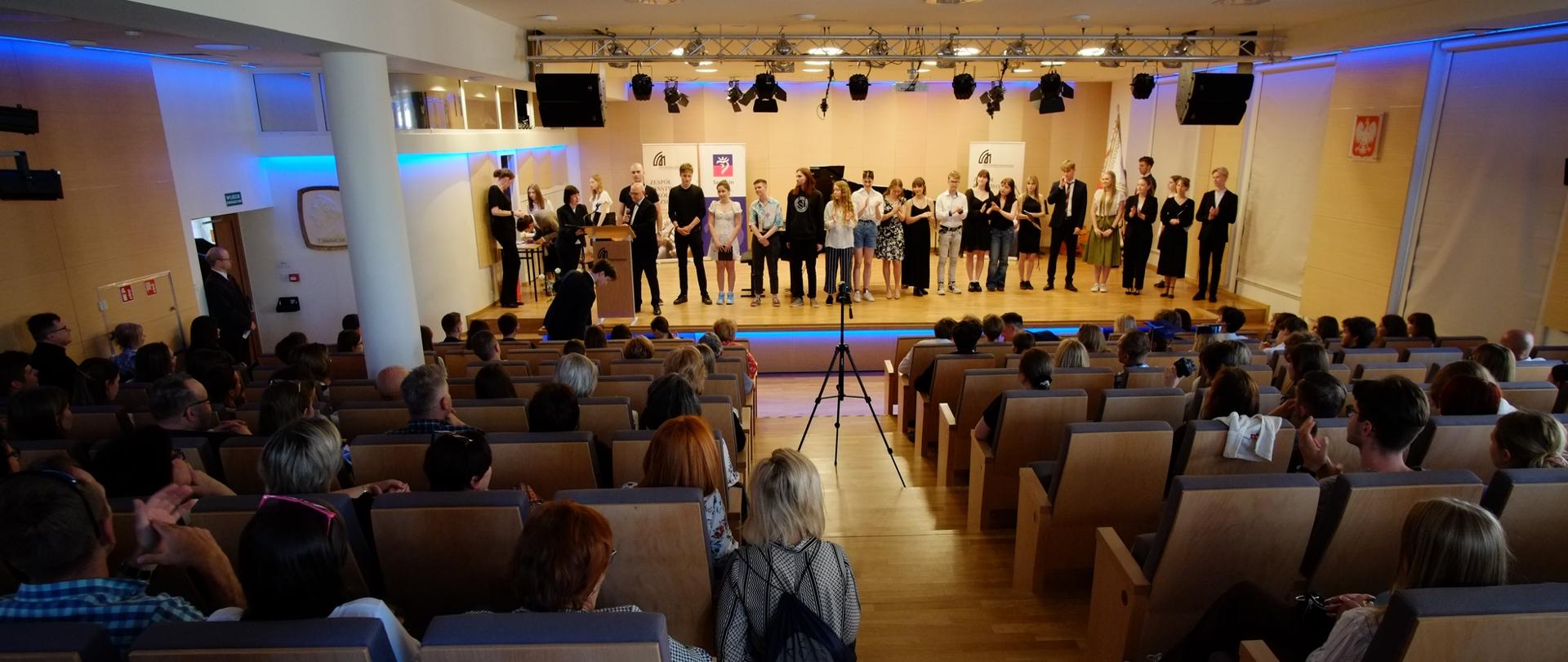 zdjęcie przedstawia maturzystów stojących w szeregu na scenie sali koncertowej, widok z tyłu widowni sali koncertowej