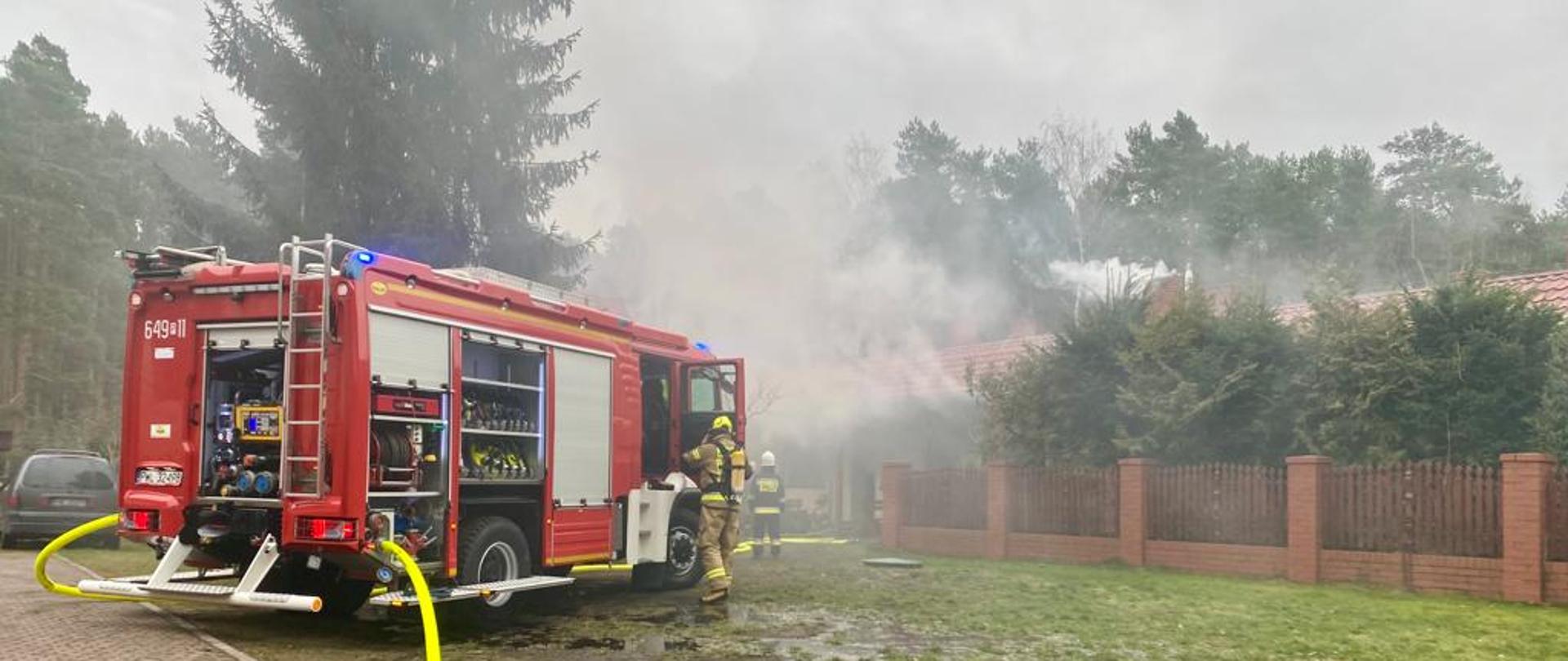 Na pierwszym planie samochód strażacki ustawiony na terenie pensjonatu ze sprawioną linią zasilającą z hydrantu, nawodnioną linią główną. Ratownik zdejmuje sprzęt ochrony okładu oddechowego po wyjściu ze strefy zadymienia. W tle obłoki dymu.