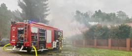 Na pierwszym planie samochód strażacki ustawiony na terenie pensjonatu ze sprawioną linią zasilającą z hydrantu, nawodnioną linią główną. Ratownik zdejmuje sprzęt ochrony okładu oddechowego po wyjściu ze strefy zadymienia. W tle obłoki dymu.