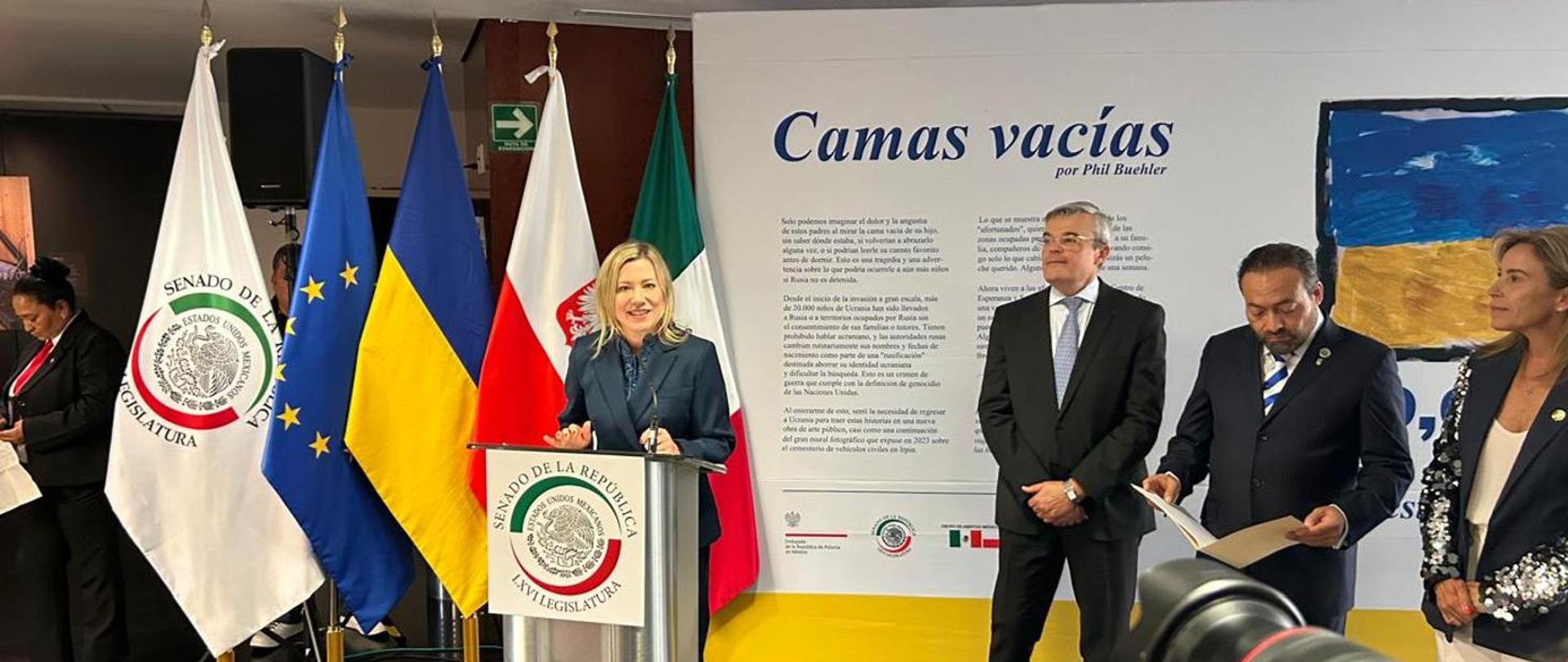 Ambasador Agnieszka Frydrychowicz-Tekieli wygłasza mowę podczas otwarcia wystawy.
W tle flagi Meksyku, Polski, Ukrainy, Unii Europejskiej i Senatu Meksyku oraz plansza wprowadzająca do wystawy „Puste łóżka".
