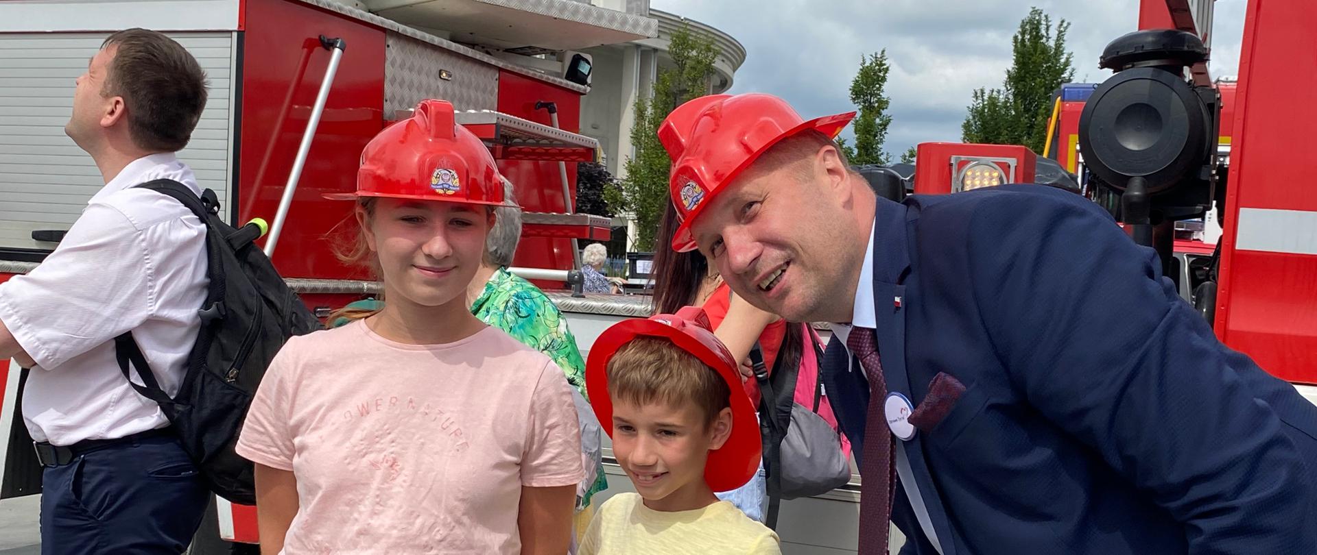 Minister robi sobie zdjęcie z uczestnikami festynu,. To dwoje dzieci. Wszyscy mają na głowach hełmy strażackie. W tle pojazdy pożarnicze.