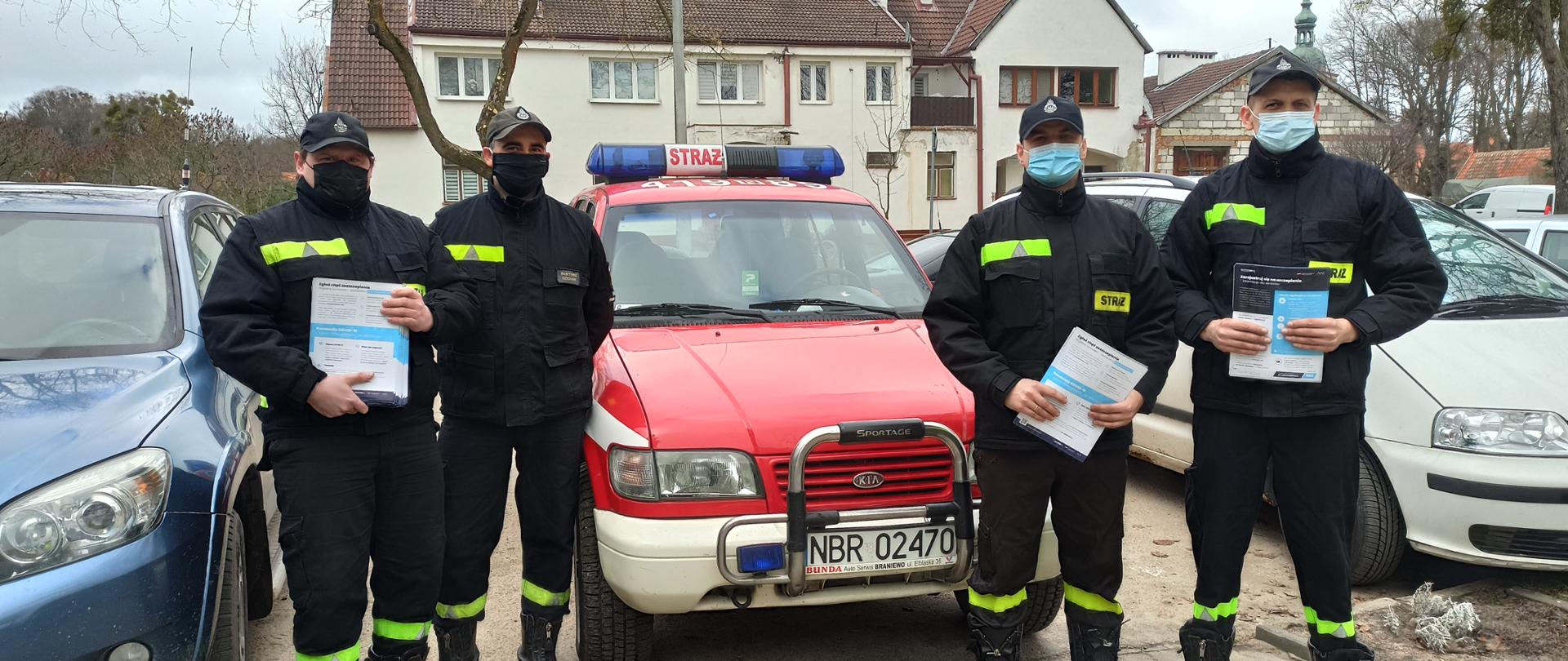 Na zdjęciu czterech strażaków w mundurach i maseczkach trzymają ulotki w rekach. Stoją przy czerwonym samochodzie strażackim. W tle inne samochody i budynek mieszkalny.