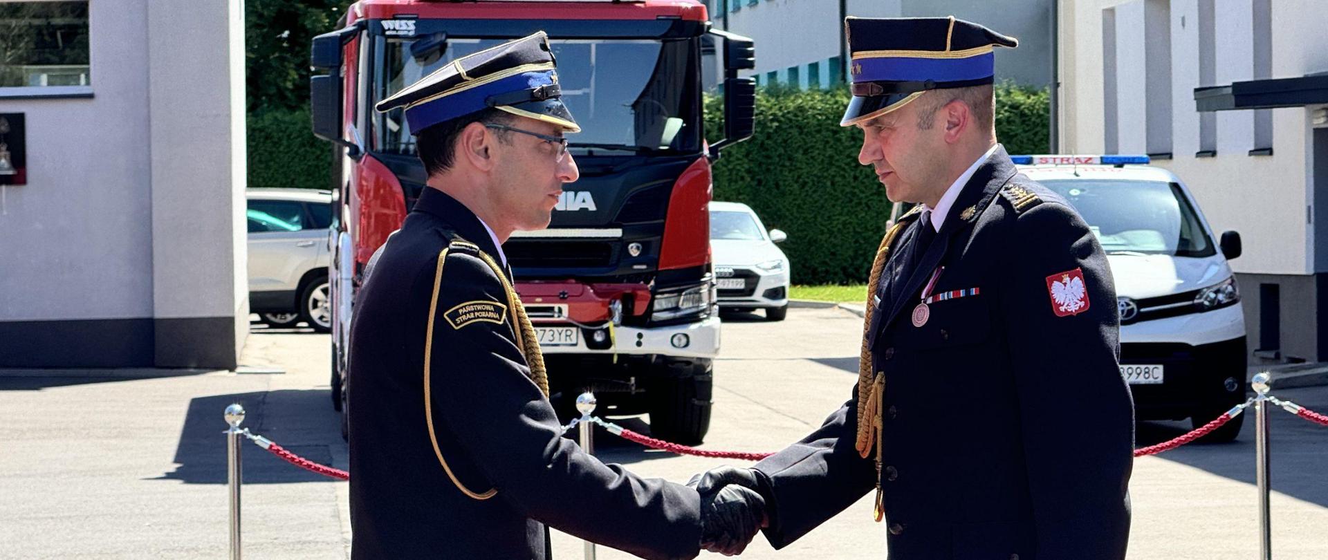 Uścisk dłoni Komendanta Wojewódzkiego st. bryg. Wojciecha Kruczka z odchodzącym na zaopatrzenie emerytalne st. bryg. Patrycjuszem Fudałą w mundurach galowych na placu wewnętrznym Komendy Miejskiej Państwowej Straży Pożarnej w Jaworznie.