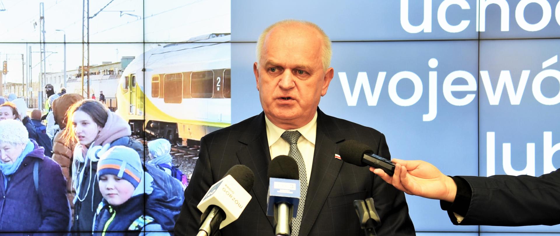 Wojewoda Władsław Dajczak w czasie briefingu przemawia przy mównicy, za plecami ma ekran z prezentacją