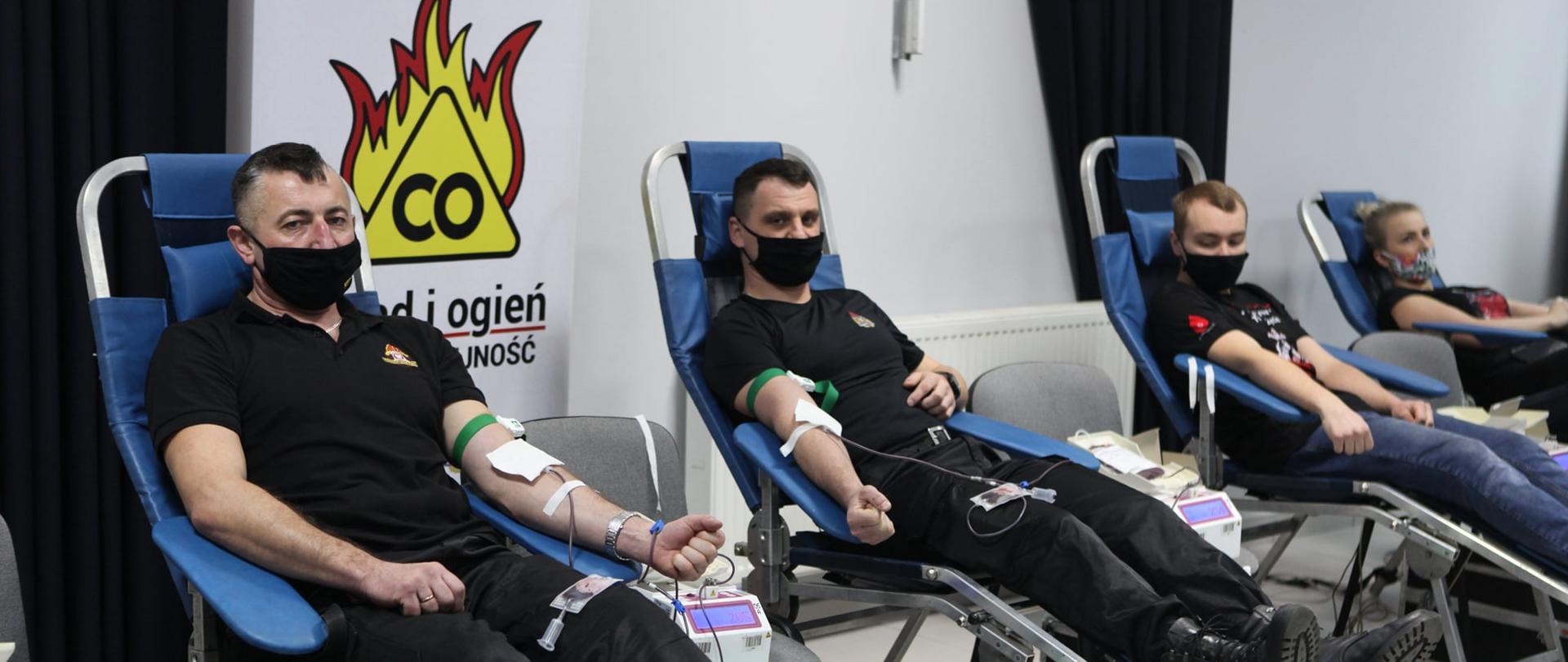 Zdjęcie przedstawia dwóch strażaków w ubraniach koszarowych oraz dwoje nastolatków, wszyscy na leżakach medycznych w trakcie oddawania krwi. W tle logo programu Czad i ogień.