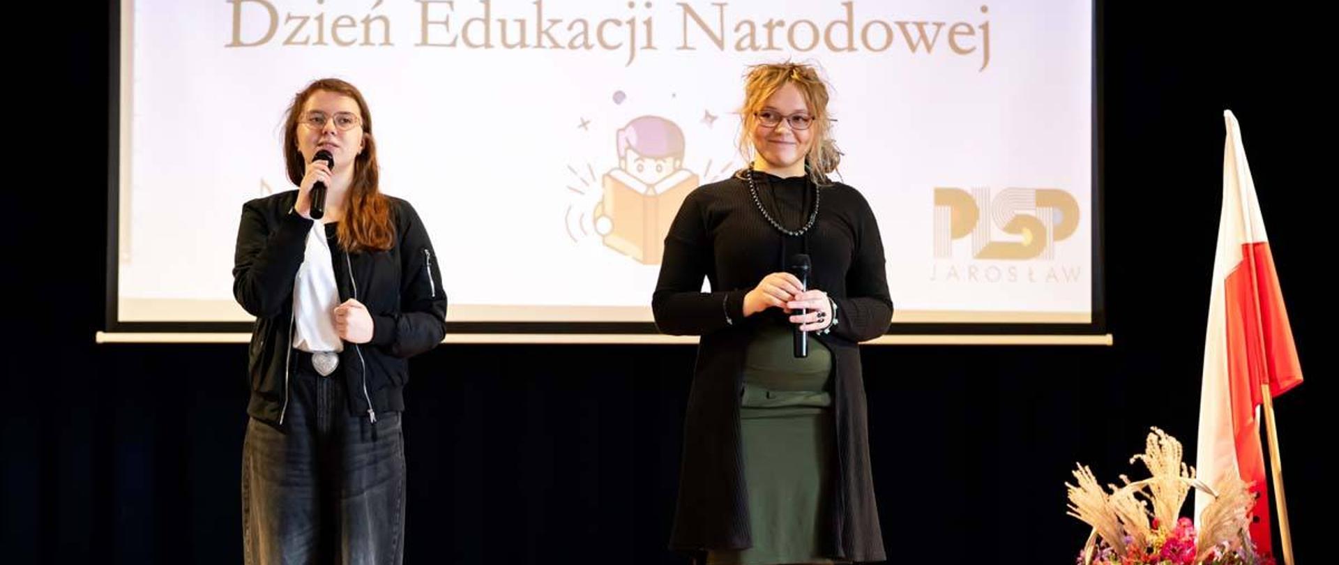 Na zdjęciu widać dwie kobiety deklamujące wiersze do mikrofonu. W tle znajduje się ekran z napisem Dzień Edukacji Narodowej.