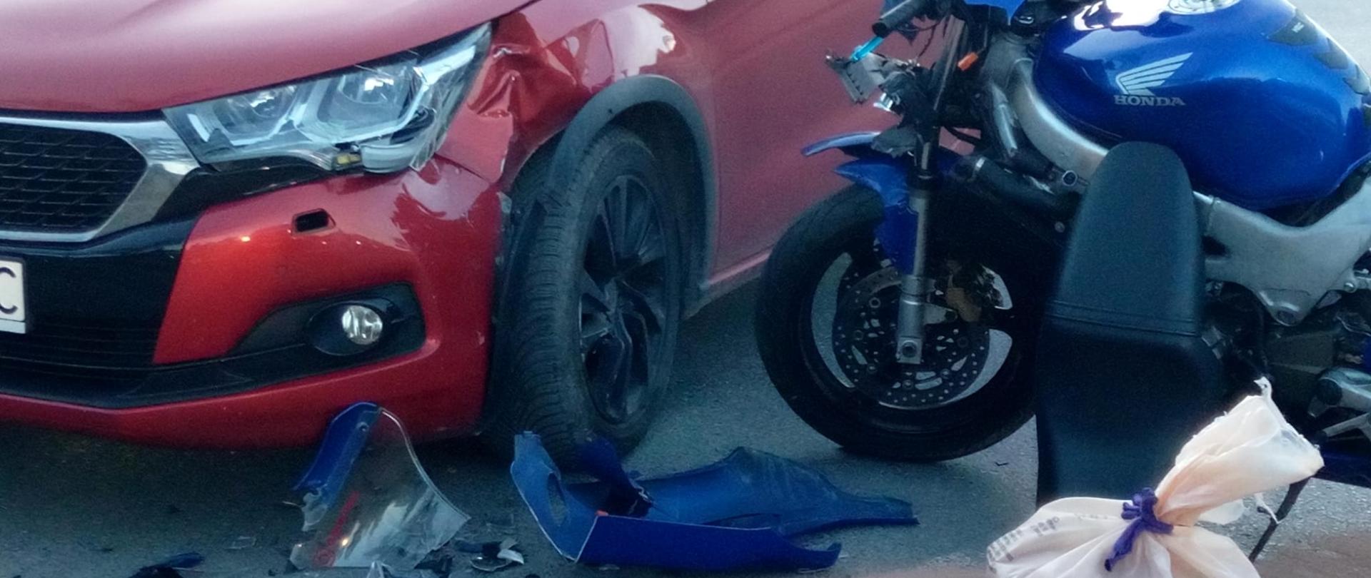 Zdjęcie przedstawia oba pojazdy biorące udział w zderzeniu. Po lewej stoi czerwony samochód osobowy zaś po prawej, prostopadle do auta motocykl Honda. Na ziemi leżą fragmenty zniszczonego jednośladu.