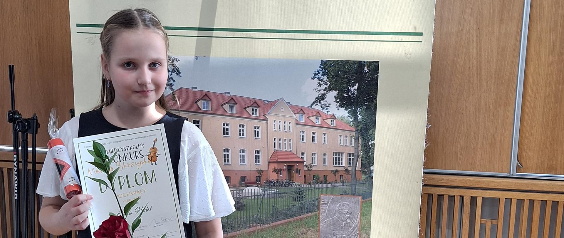 Uczennica jest elegancko ubrana. Stoi przed banerem reklamującym Państwową Szkołę Muzyczną w Lesznie. W rękach trzyma różę, dyplom i nagrodę. 
