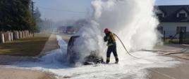 Na zdjęciu widzimy działania gaśnicze przy palącym się samochodzie. Strażacy zabezpieczeni w aparaty ochrony dróg oddechowych gaszą pojazd. W tle widać wozy strażackie.