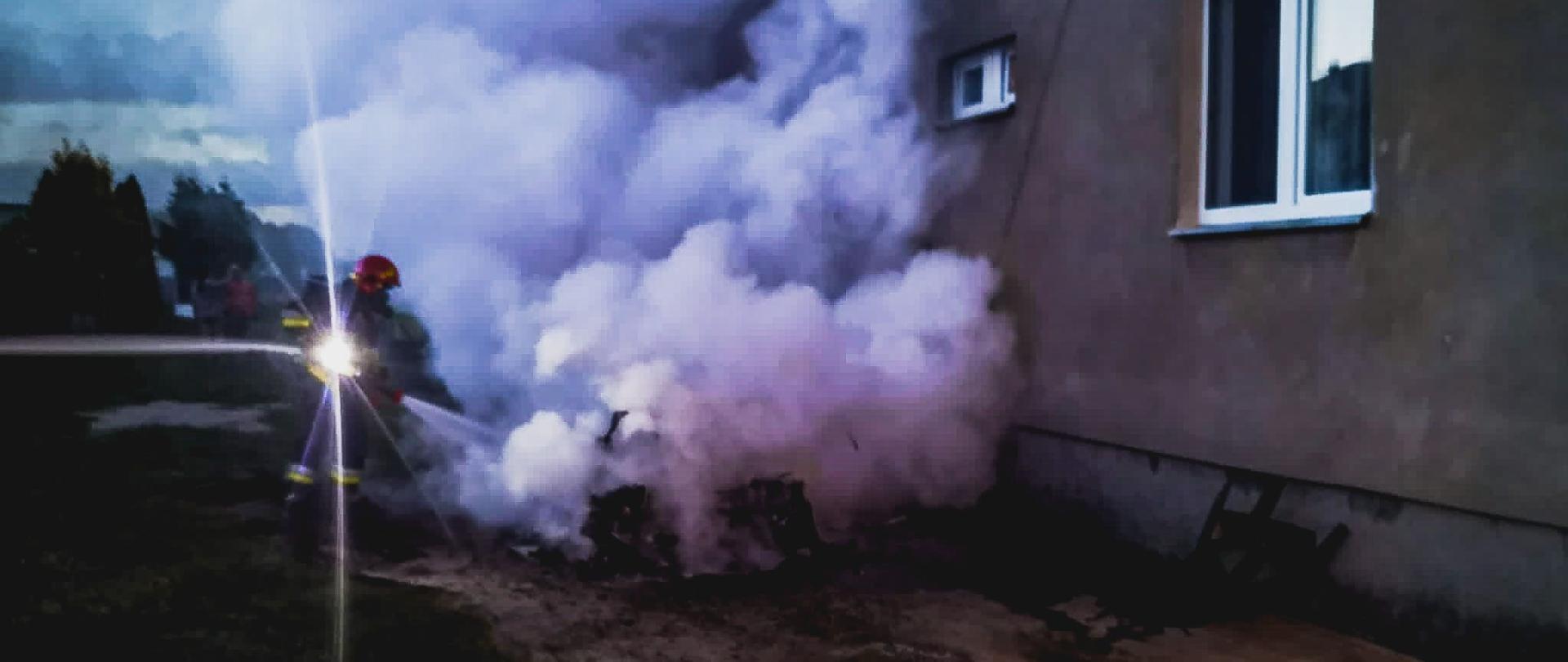 Strażak w ubraniu i w aparacie powietrznym gasi pożar samochodu, z którego wydobywają się kłęby dymu oraz pary. Po prawej stronie zagrożony budynek mieszkalny.