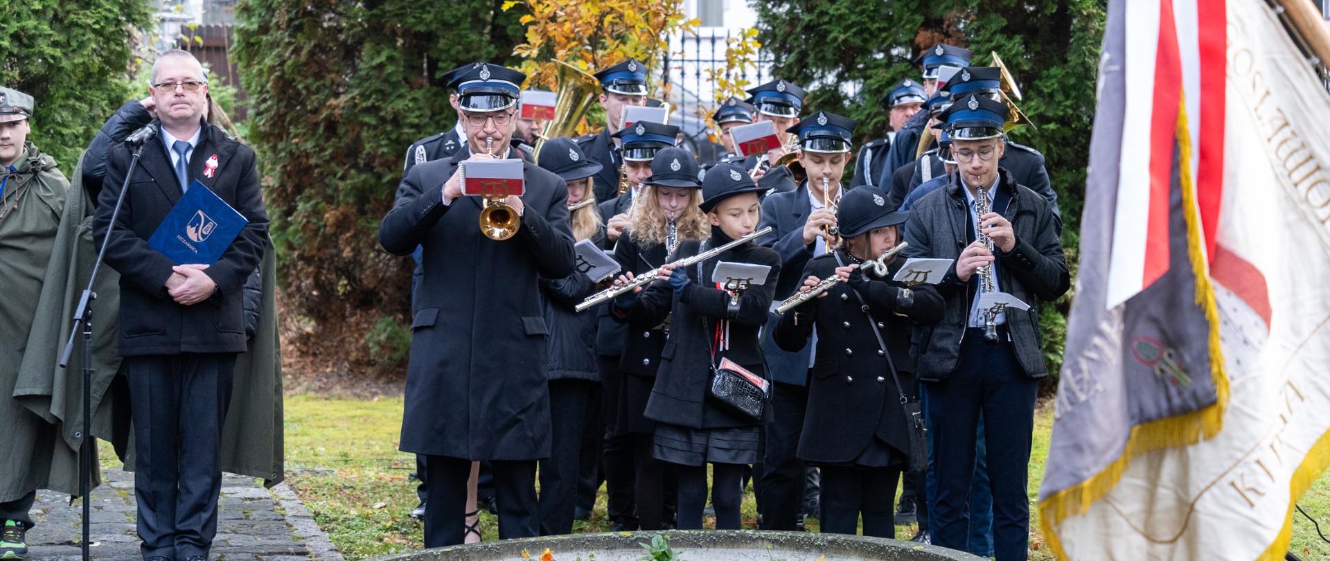 Zdjęcie w plenerze. Orkiestra dęta złożona między innymi z uczniów filii Jaworki. Z prawej strony sztandar. Na środku uczestnicy orkiestry grają na instrumentach dętych blaszanych i drewnianych. Za nimi drzewa liściaste oraz iglaste, a w tle okna budynku mieszkalnego. Z lewej strony mikrofon, przed którym stoi osoba trzymająca teczkę z logo miasta. Za nim stoją harcerze. Na pierwszym planie okrągły kwietnik, a w nim posadzone bratki