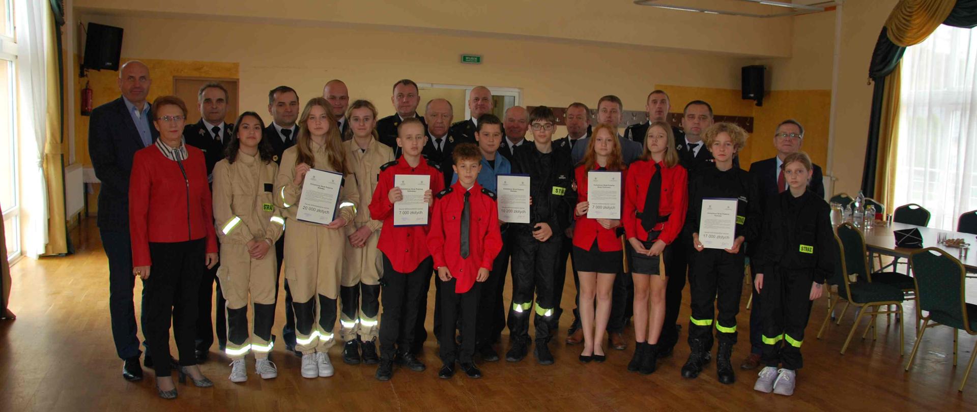 W pomieszczeniu na dużej Sali stoi kilkanaście osób w tym strażacy PSP, OSP i osoby cywilne. Przed nimi stoi kilkanaście dzieci, część z nich prezentuje dokumenty.
